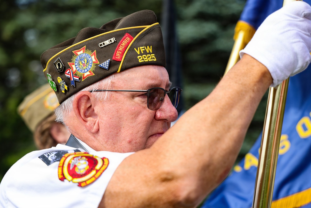 An older male veteran is participating in a formal color guard or ceremonial event, holding a flag staff while wearing a VFW (Veterans of Foreign Wars) garrison cap with multiple pins and patches. His cap identifies him as a member of VFW Post 2925 and a Life Member, with a 'COMDR' (Commander) designation visible. He is dressed in a white shirt with a Marine Corps emblem on the sleeve and is wearing white ceremonial gloves.