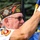 An older male veteran is participating in a formal color guard or ceremonial event, holding a flag staff while wearing a VFW (Veterans of Foreign Wars) garrison cap with multiple pins and patches. His cap identifies him as a member of VFW Post 2925 and a Life Member, with a 'COMDR' (Commander) designation visible. He is dressed in a white shirt with a Marine Corps emblem on the sleeve and is wearing white ceremonial gloves.