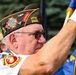 An older male veteran is participating in a formal color guard or ceremonial event, holding a flag staff while wearing a VFW (Veterans of Foreign Wars) garrison cap with multiple pins and patches. His cap identifies him as a member of VFW Post 2925 and a Life Member, with a 'COMDR' (Commander) designation visible. He is dressed in a white shirt with a Marine Corps emblem on the sleeve and is wearing white ceremonial gloves.