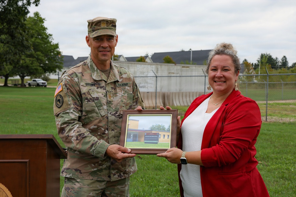 A U.S. Army soldier identified by name tape as 'BROWN' and a civilian woman in a red blazer jointly hold a framed photograph or plaque during what appears to be an outdoor ceremony. The soldier is wearing Army Combat Uniform (ACU) in OCP (Operational Camouflage Pattern) with U.S. Army insignia and an American flag patch. The setting appears to be on a military installation or base grounds.