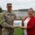 A U.S. Army soldier identified by name tape as 'BROWN' and a civilian woman in a red blazer jointly hold a framed photograph or plaque during what appears to be an outdoor ceremony. The soldier is wearing Army Combat Uniform (ACU) in OCP (Operational Camouflage Pattern) with U.S. Army insignia and an American flag patch. The setting appears to be on a military installation or base grounds.