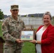 A U.S. Army soldier identified by name tape as 'BROWN' and a civilian woman in a red blazer jointly hold a framed photograph or plaque during what appears to be an outdoor ceremony. The soldier is wearing Army Combat Uniform (ACU) in OCP (Operational Camouflage Pattern) with U.S. Army insignia and an American flag patch. The setting appears to be on a military installation or base grounds.