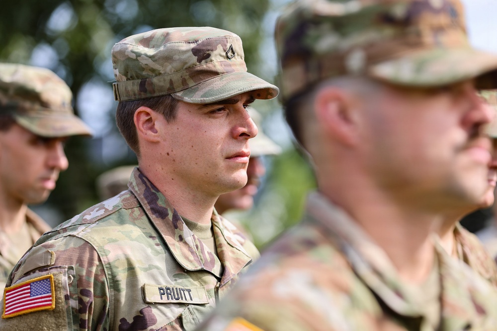The image shows multiple U.S. Army soldiers in Operational Camouflage Pattern (OCP) uniforms standing in formation. The central figure, identified by a name tape as 'PRUITT,' is in sharp focus and appears to be at attention or participating in a formal military ceremony or formation. An American flag patch is visible on the left shoulder of the central soldier.