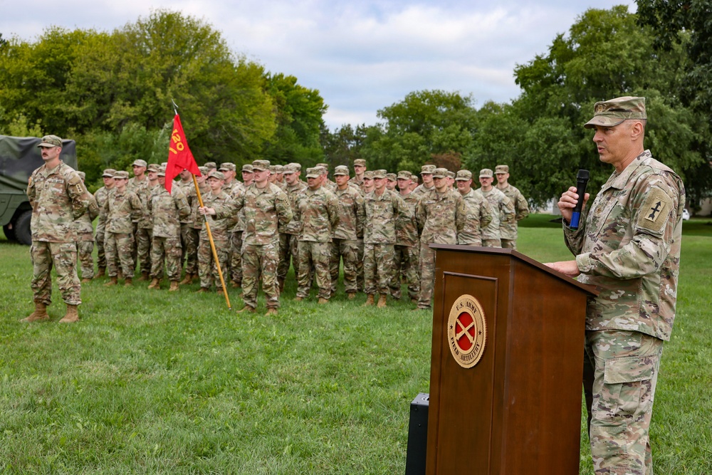 A U.S. Army Field Artillery ceremony is taking place outdoors on a grass field. A senior officer in OCP (Operational Camouflage Pattern) uniform is speaking at a wooden podium bearing the U.S. Army Field Artillery seal, while a formation of soldiers stands at attention in the background. A guidon bearer holds a red unit flag with yellow markings to the left of the formation.