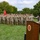 A U.S. Army Field Artillery ceremony is taking place outdoors on a grass field. A senior officer in OCP (Operational Camouflage Pattern) uniform is speaking at a wooden podium bearing the U.S. Army Field Artillery seal, while a formation of soldiers stands at attention in the background. A guidon bearer holds a red unit flag with yellow markings to the left of the formation.