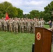 A U.S. Army Field Artillery ceremony is taking place outdoors on a grass field. A senior officer in OCP (Operational Camouflage Pattern) uniform is speaking at a wooden podium bearing the U.S. Army Field Artillery seal, while a formation of soldiers stands at attention in the background. A guidon bearer holds a red unit flag with yellow markings to the left of the formation.