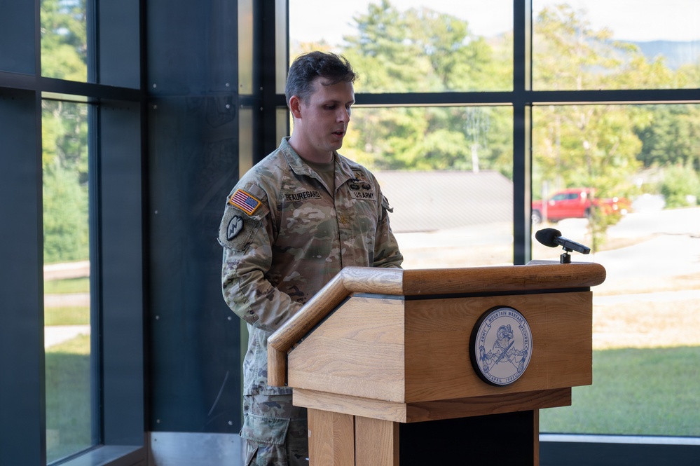 A U.S. Army soldier with the name tape 'BEAUREGARD' is speaking at a wooden podium inside a modern building with large windows. The podium bears a seal that appears to read 'Army Mountain Warfare School.' The setting suggests an official military ceremony or briefing.