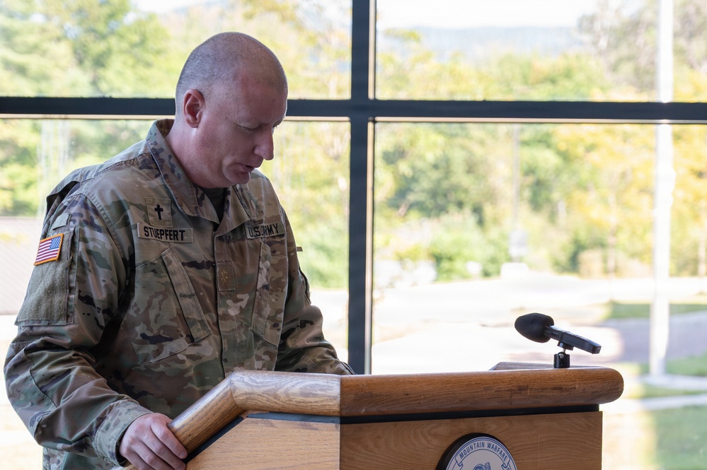 A U.S. Army chaplain identified by name tape as 'STUEPFERT' stands at a wooden podium, appearing to read or pray with head bowed. He is wearing the OCP (Operational Camouflage Pattern) uniform with a chaplain's cross insignia and a U.S. flag patch on his left shoulder. The podium bears a seal that reads 'Mountain Warfare,' suggesting a military installation or training center setting.