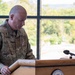 A U.S. Army chaplain identified by name tape as 'STUEPFERT' stands at a wooden podium, appearing to read or pray with head bowed. He is wearing the OCP (Operational Camouflage Pattern) uniform with a chaplain's cross insignia and a U.S. flag patch on his left shoulder. The podium bears a seal that reads 'Mountain Warfare,' suggesting a military installation or training center setting.
