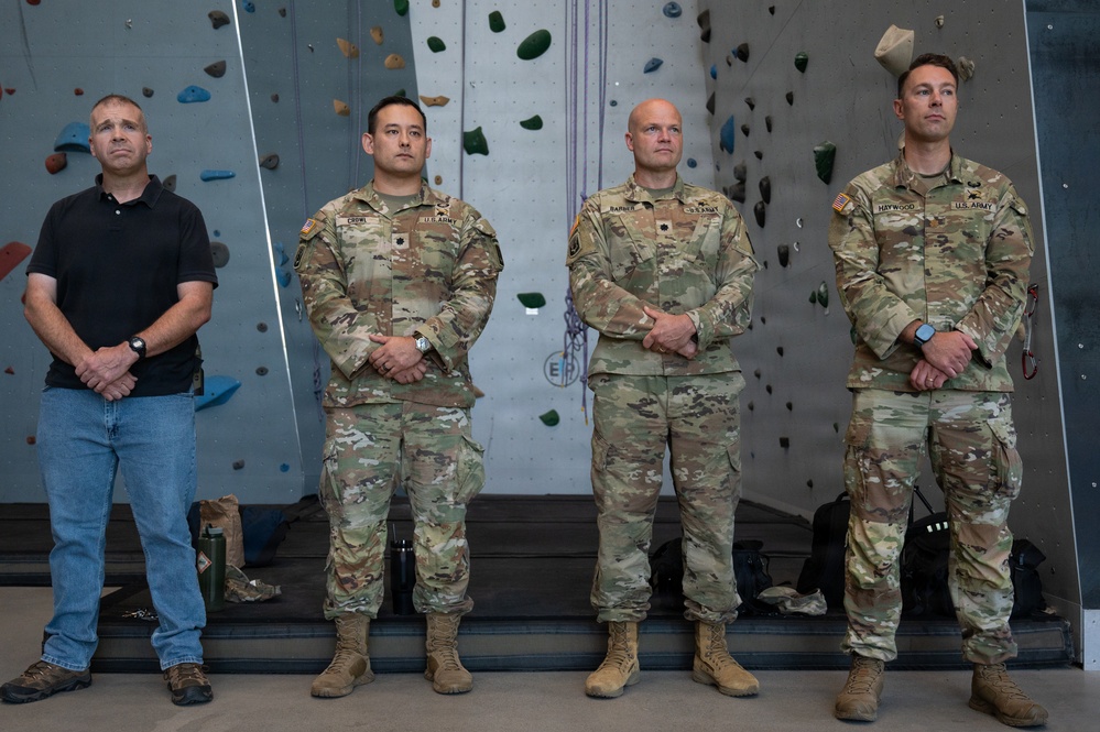 Four men stand at attention in front of an indoor rock climbing wall. Three are wearing U.S. Army OCP (Operational Camouflage Pattern) uniforms with visible name tapes, while one man on the left is in civilian attire (black polo shirt and jeans). The setting appears to be a formal or ceremonial occasion at a military or recreational facility.