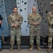 Four men stand at attention in front of an indoor rock climbing wall. Three are wearing U.S. Army OCP (Operational Camouflage Pattern) uniforms with visible name tapes, while one man on the left is in civilian attire (black polo shirt and jeans). The setting appears to be a formal or ceremonial occasion at a military or recreational facility.