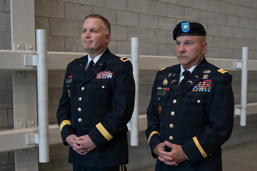 Two U.S. Army officers in dress blue uniforms stand at attention in what appears to be an indoor military facility. Both wear extensive medal and ribbon arrays indicating significant service records. The officer on the right wears a black beret with a distinctive blue and silver flash, while the officer on the left appears bare-headed.