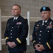 Two U.S. Army officers in dress blue uniforms stand at attention in what appears to be an indoor military facility. Both wear extensive medal and ribbon arrays indicating significant service records. The officer on the right wears a black beret with a distinctive blue and silver flash, while the officer on the left appears bare-headed.