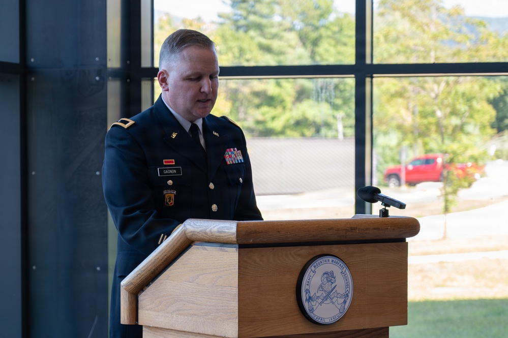 A U.S. Army soldier in dress blues uniform, identified by nameplate as 'Gagnon', is speaking at a wooden podium bearing the seal of the Army Mountain Warfare School. The setting appears to be an official military ceremony or formal address inside a modern building with large windows overlooking a wooded outdoor area. The soldier displays several service ribbons and unit patches on their uniform.