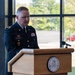 A U.S. Army soldier in dress blues uniform, identified by nameplate as 'Gagnon', is speaking at a wooden podium bearing the seal of the Army Mountain Warfare School. The setting appears to be an official military ceremony or formal address inside a modern building with large windows overlooking a wooded outdoor area. The soldier displays several service ribbons and unit patches on their uniform.