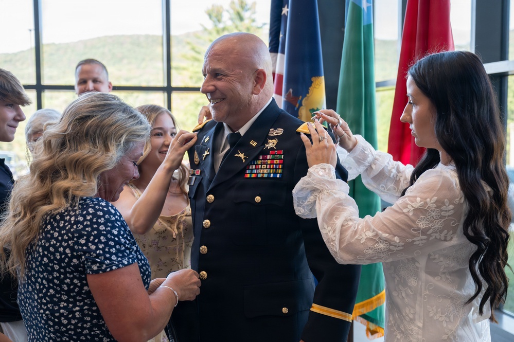A U.S. Army officer in dress blues is having rank insignia pinned onto his shoulders by two women, likely family members, during a military promotion ceremony. The officer is smiling broadly while the women attach gold rank devices to his epaulettes. Multiple flags and additional attendees are visible in the background of what appears to be an indoor ceremonial venue.