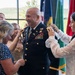 A U.S. Army officer in dress blues is having rank insignia pinned onto his shoulders by two women, likely family members, during a military promotion ceremony. The officer is smiling broadly while the women attach gold rank devices to his epaulettes. Multiple flags and additional attendees are visible in the background of what appears to be an indoor ceremonial venue.