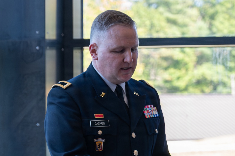 A U.S. Army soldier in dress blues uniform is photographed indoors near a large window with trees visible in the background. The soldier's name tape reads 'GAGNON' and displays multiple service ribbons and insignia. He appears to be looking downward, possibly reading or speaking at a formal event.