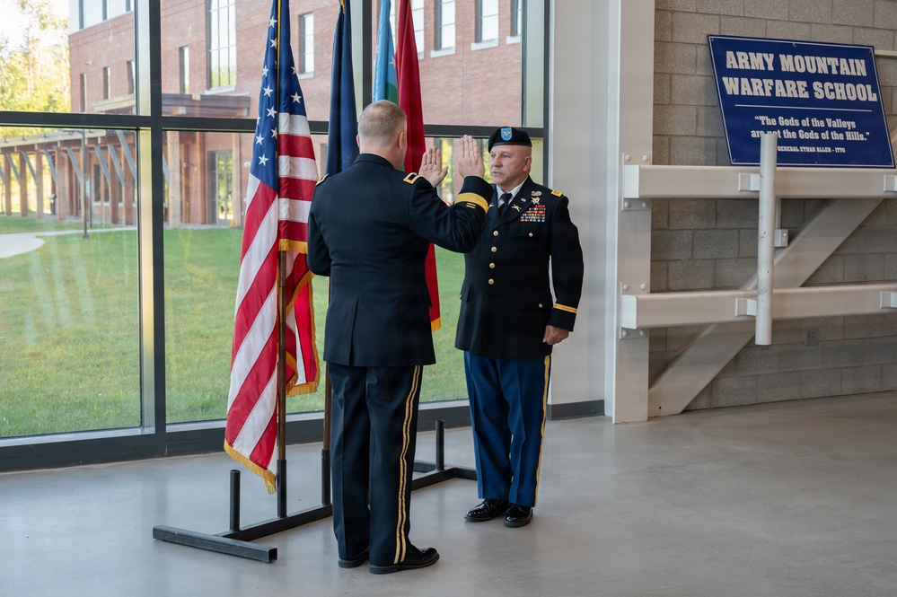 Two U.S. Army officers in dress uniforms are conducting a formal oath or promotion ceremony at the Army Mountain Warfare School. Both individuals have their right hands raised, indicating an oath of office or commissioning ceremony. The setting is an indoor facility with an American flag and other military flags displayed prominently.