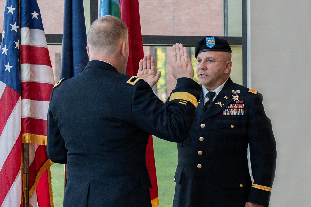 Two U.S. Army officers in dress blue uniforms are participating in a formal oath of office or promotion ceremony, with both raising their right hands. The officer facing the camera wears a black beret with a flash, multiple service ribbons, and general officer rank insignia. The ceremony takes place indoors in front of multiple flags including the U.S. flag and Army flag.