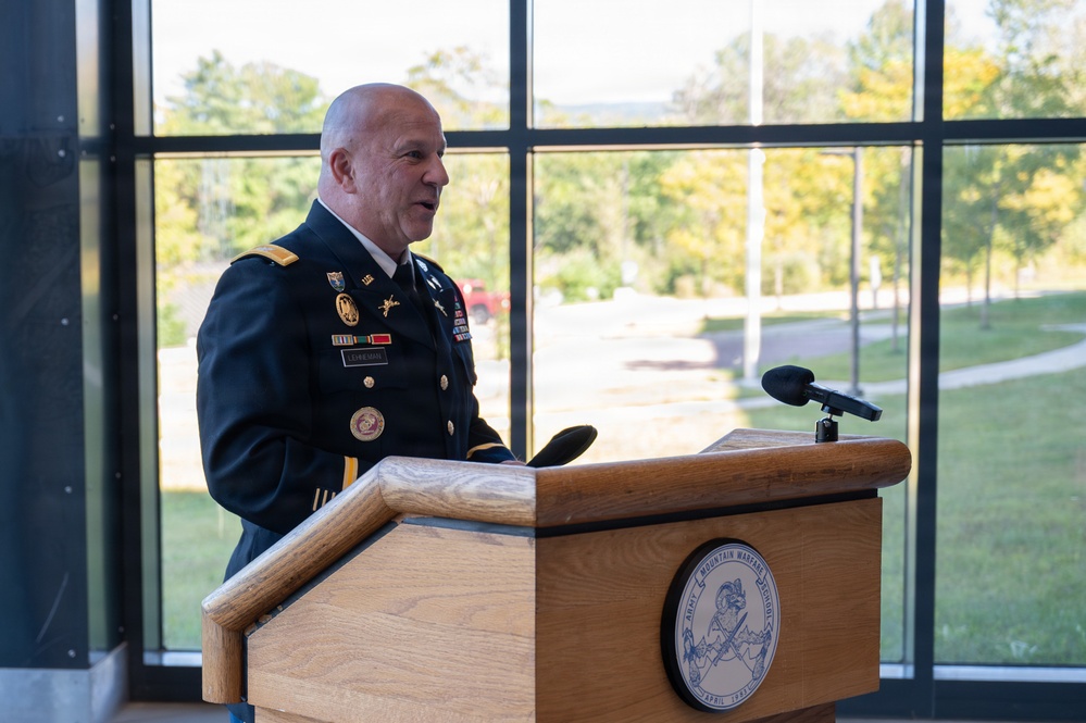 A U.S. Army officer in dress blues uniform is speaking at a wooden podium bearing the seal of the Army Mountain Warfare School. The setting appears to be an indoor facility with large windows overlooking a green outdoor area. The officer displays multiple medals, ribbons, and rank insignia consistent with a senior officer.