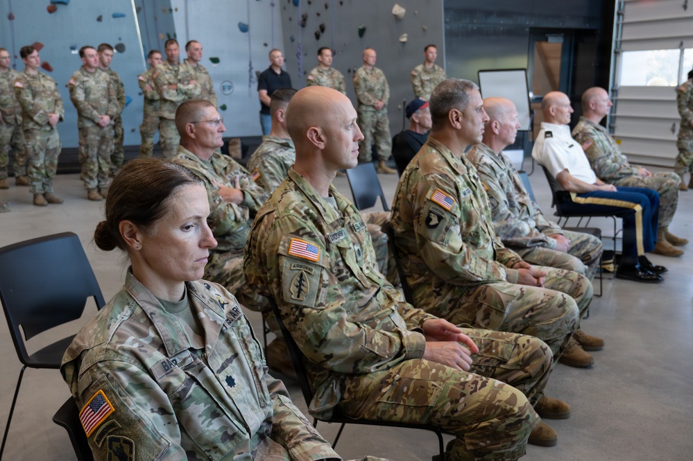 A group of U.S. Army soldiers in OCP (Operational Camouflage Pattern) uniforms are gathered inside what appears to be a military fitness facility with a climbing wall visible in the background. Senior personnel are seated in the foreground while additional soldiers stand in formation behind them, suggesting a briefing, ceremony, or official event. One individual in the background is wearing a white dress uniform shirt, indicating a mix of ranks and possibly a formal occasion.