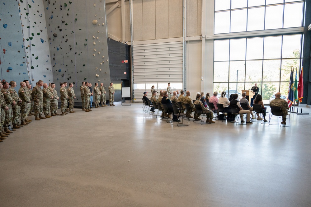 A military ceremony is taking place inside a large indoor facility featuring a prominent rock climbing wall on the left side. A formation of uniformed military personnel stands at attention along the left wall while seated attendees, including both military and civilian individuals, face a speaker at a podium on the right side. Flags are visible near the podium, indicating a formal official event.