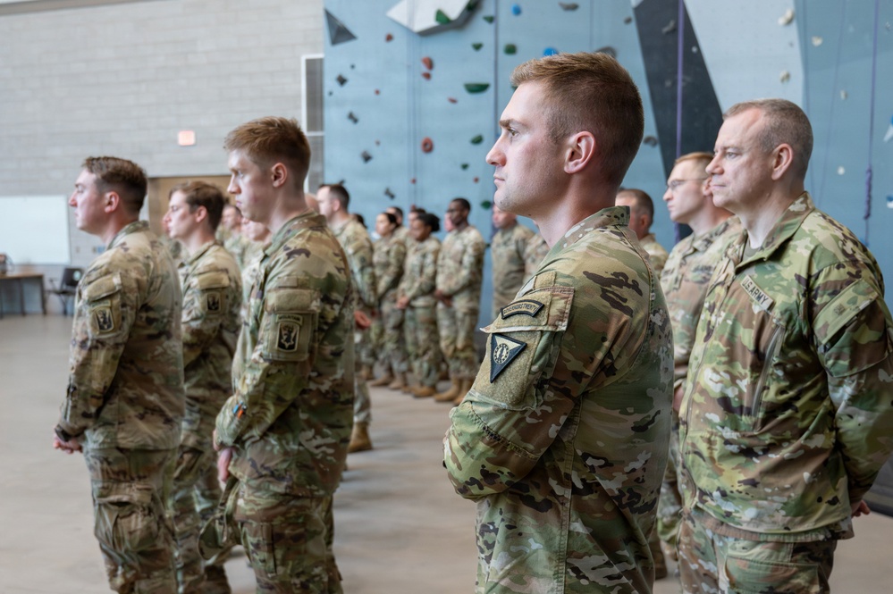 A formation of U.S. Army soldiers in OCP (Operational Camouflage Pattern) uniforms standing at attention inside an indoor facility with a rock climbing wall visible in the background. The soldiers appear to be participating in a formal military ceremony or briefing. Multiple rows of personnel are visible, with varying ranks indicated by shoulder patches and insignia.