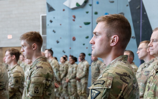 A formation of U.S. Army soldiers in OCP (Operational Camouflage Pattern) uniforms standing at attention inside an indoor facility with a rock climbing wall visible in the background. The soldiers appear to be participating in a formal military ceremony or briefing. Multiple rows of personnel are visible, with varying ranks indicated by shoulder patches and insignia.