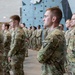 A formation of U.S. Army soldiers in OCP (Operational Camouflage Pattern) uniforms standing at attention inside an indoor facility with a rock climbing wall visible in the background. The soldiers appear to be participating in a formal military ceremony or briefing. Multiple rows of personnel are visible, with varying ranks indicated by shoulder patches and insignia.