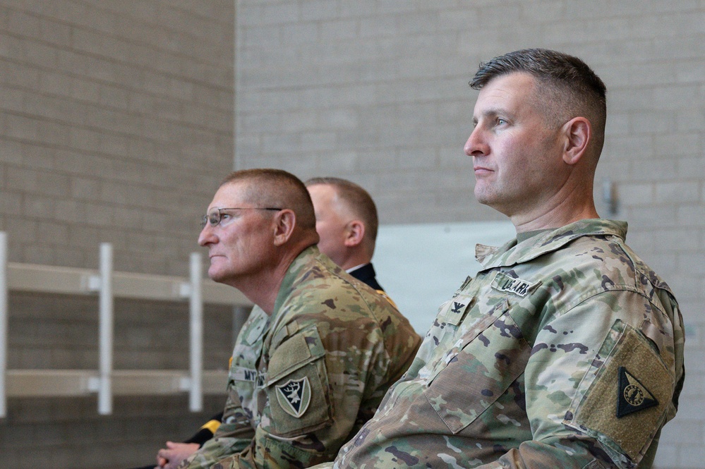 Three military personnel in U.S. Army OCP (Operational Camouflage Pattern) uniforms are seated and facing the same direction, appearing to observe an event or ceremony. Two individuals are clearly visible in the foreground, with a third partially visible in the background. The setting appears to be an indoor facility with concrete block walls.