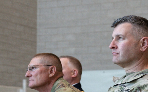 Three military personnel in U.S. Army OCP (Operational Camouflage Pattern) uniforms are seated and facing the same direction, appearing to observe an event or ceremony. Two individuals are clearly visible in the foreground, with a third partially visible in the background. The setting appears to be an indoor facility with concrete block walls.
