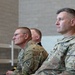 Three military personnel in U.S. Army OCP (Operational Camouflage Pattern) uniforms are seated and facing the same direction, appearing to observe an event or ceremony. Two individuals are clearly visible in the foreground, with a third partially visible in the background. The setting appears to be an indoor facility with concrete block walls.
