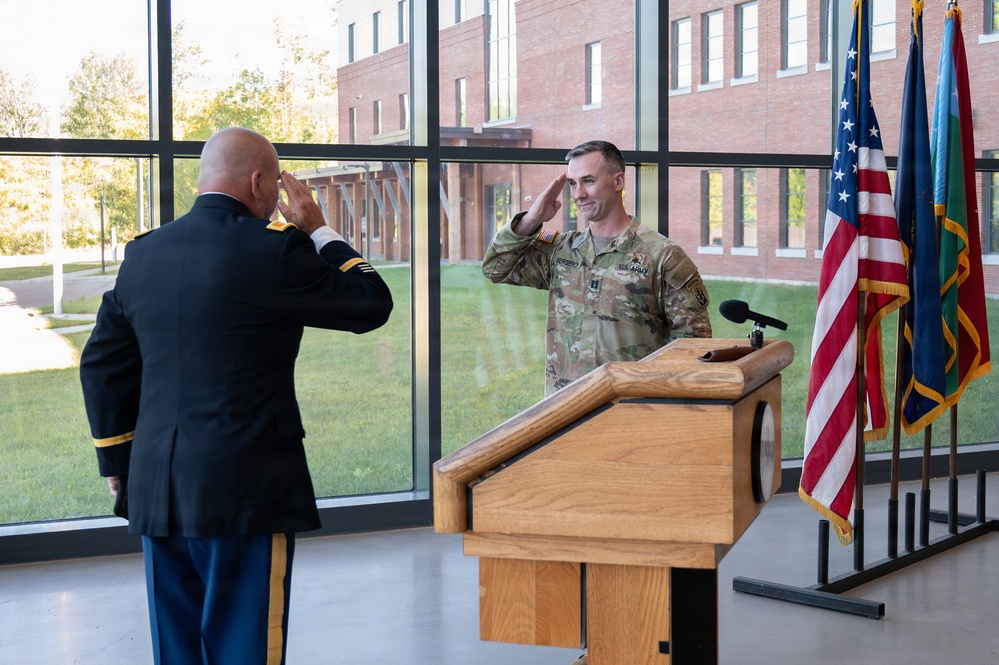 Two U.S. Army soldiers are exchanging salutes during what appears to be a formal military ceremony, likely a promotion or change of command. One soldier is in dress blues uniform facing away from the camera, while the other is in Army Combat Uniform (ACU/OCP) standing at a wooden podium. The event is taking place indoors in a modern facility with large windows overlooking a brick building exterior.