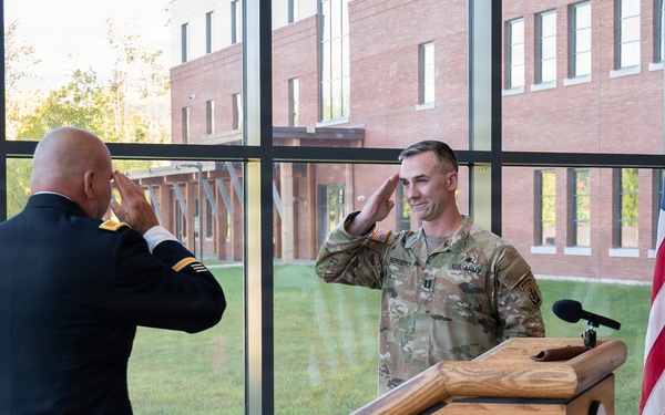 Two U.S. Army soldiers are exchanging salutes during what appears to be a formal military ceremony, likely a promotion or change of command. One soldier is in dress blues uniform facing away from the camera, while the other is in Army Combat Uniform (ACU/OCP) standing at a wooden podium. The event is taking place indoors in a modern facility with large windows overlooking a brick building exterior.
