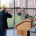 Two U.S. Army soldiers are exchanging salutes during what appears to be a formal military ceremony, likely a promotion or change of command. One soldier is in dress blues uniform facing away from the camera, while the other is in Army Combat Uniform (ACU/OCP) standing at a wooden podium. The event is taking place indoors in a modern facility with large windows overlooking a brick building exterior.