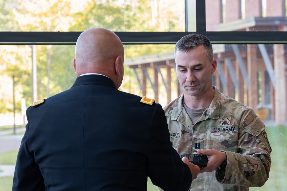 Two U.S. Army soldiers are engaged in what appears to be a pinning or award ceremony indoors near a large window. One soldier is in Army Service Uniform (dress blues) with his back to the camera, while the other in Army Combat Uniform (OCP/multicam) is holding a small object, likely a rank insignia or medal. The name tape on the soldier in OCP reads 'ERON' (partially visible, possibly 'CAMERON' or similar).