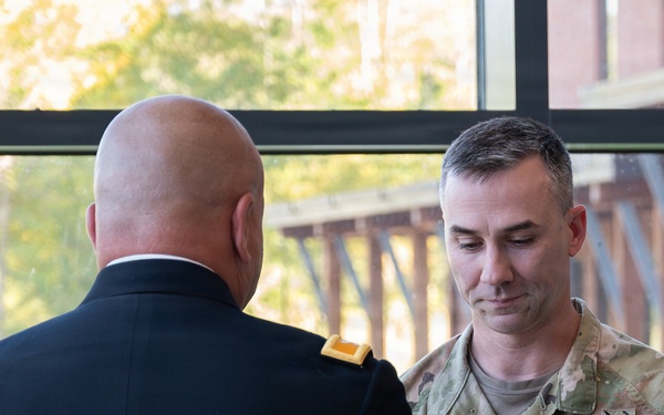 Two U.S. Army soldiers are engaged in what appears to be a pinning or award ceremony indoors near a large window. One soldier is in Army Service Uniform (dress blues) with his back to the camera, while the other in Army Combat Uniform (OCP/multicam) is holding a small object, likely a rank insignia or medal. The name tape on the soldier in OCP reads 'ERON' (partially visible, possibly 'CAMERON' or similar).
