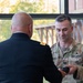 Two U.S. Army soldiers are engaged in what appears to be a pinning or award ceremony indoors near a large window. One soldier is in Army Service Uniform (dress blues) with his back to the camera, while the other in Army Combat Uniform (OCP/multicam) is holding a small object, likely a rank insignia or medal. The name tape on the soldier in OCP reads 'ERON' (partially visible, possibly 'CAMERON' or similar).