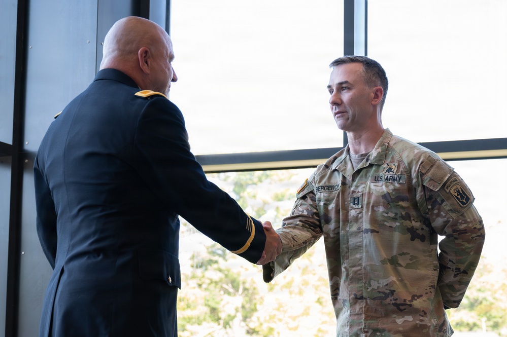 Two U.S. Army personnel are shaking hands in what appears to be an official or ceremonial setting. One individual is wearing a formal Army Service Uniform (ASU) in dark blue, while the other is wearing Operational Camouflage Pattern (OCP) combat uniform with the name tape 'BERGERON' and U.S. Army tape visible. The setting appears to be an indoor facility with large windows in the background.