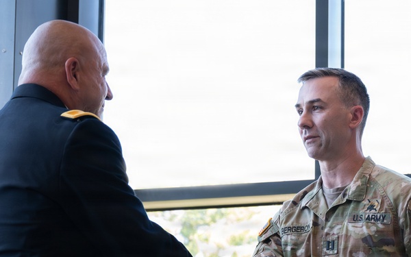 Two U.S. Army personnel are shaking hands in what appears to be an official or ceremonial setting. One individual is wearing a formal Army Service Uniform (ASU) in dark blue, while the other is wearing Operational Camouflage Pattern (OCP) combat uniform with the name tape 'BERGERON' and U.S. Army tape visible. The setting appears to be an indoor facility with large windows in the background.