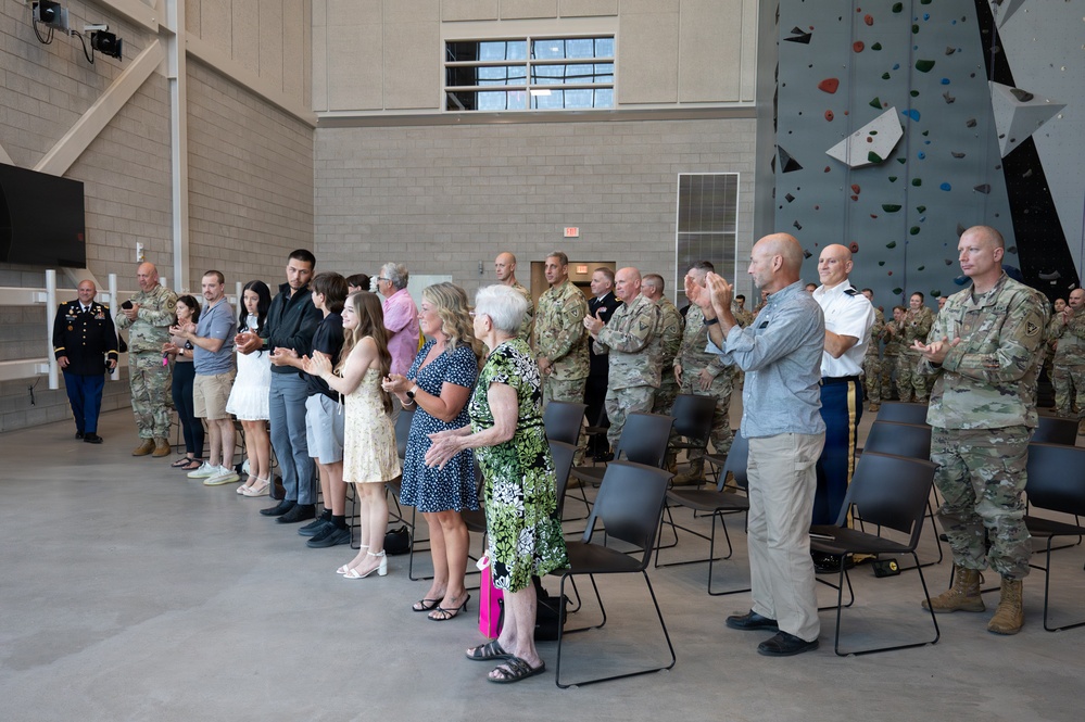 A military ceremony is taking place inside a large indoor facility featuring a rock climbing wall in the background. A mixed group of military personnel in camouflage uniforms and civilians, including family members, are standing and applauding. The event appears to be a formal recognition or promotion ceremony based on the organized standing arrangement and dress uniforms present.