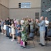 A military ceremony is taking place inside a large indoor facility featuring a rock climbing wall in the background. A mixed group of military personnel in camouflage uniforms and civilians, including family members, are standing and applauding. The event appears to be a formal recognition or promotion ceremony based on the organized standing arrangement and dress uniforms present.