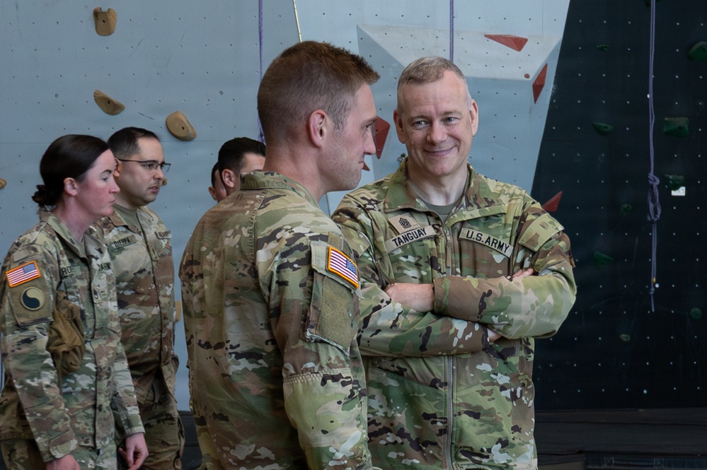 A group of U.S. Army soldiers in OCP (Operational Camouflage Pattern) uniforms are gathered inside what appears to be a military fitness facility with a rock climbing wall in the background. The soldier on the right, identified by his name tape as 'TANGUAY,' stands with arms crossed and is smiling while engaged in conversation. Several other soldiers are visible in the background, including individuals with name tapes 'ROS__' and 'DIERPA.'