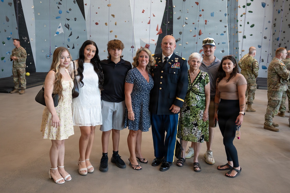 A group of eight people pose for a family photo in front of an indoor rock climbing wall. One individual is wearing a U.S. Army dress uniform with medals and ribbons, suggesting a military ceremony or promotion event. Other military personnel in combat uniforms are visible in the background.