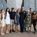 A group of eight people pose for a family photo in front of an indoor rock climbing wall. One individual is wearing a U.S. Army dress uniform with medals and ribbons, suggesting a military ceremony or promotion event. Other military personnel in combat uniforms are visible in the background.