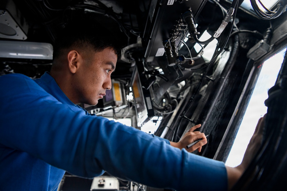 A young man wearing a blue long-sleeve shirt is performing maintenance or inspection work on complex mechanical and electrical equipment. He appears to be inside a large vehicle or aircraft, working on an overhead or side-mounted system with cables, wiring, and mechanical components. The setting suggests a military or aviation maintenance environment.