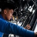A young man wearing a blue long-sleeve shirt is performing maintenance or inspection work on complex mechanical and electrical equipment. He appears to be inside a large vehicle or aircraft, working on an overhead or side-mounted system with cables, wiring, and mechanical components. The setting suggests a military or aviation maintenance environment.