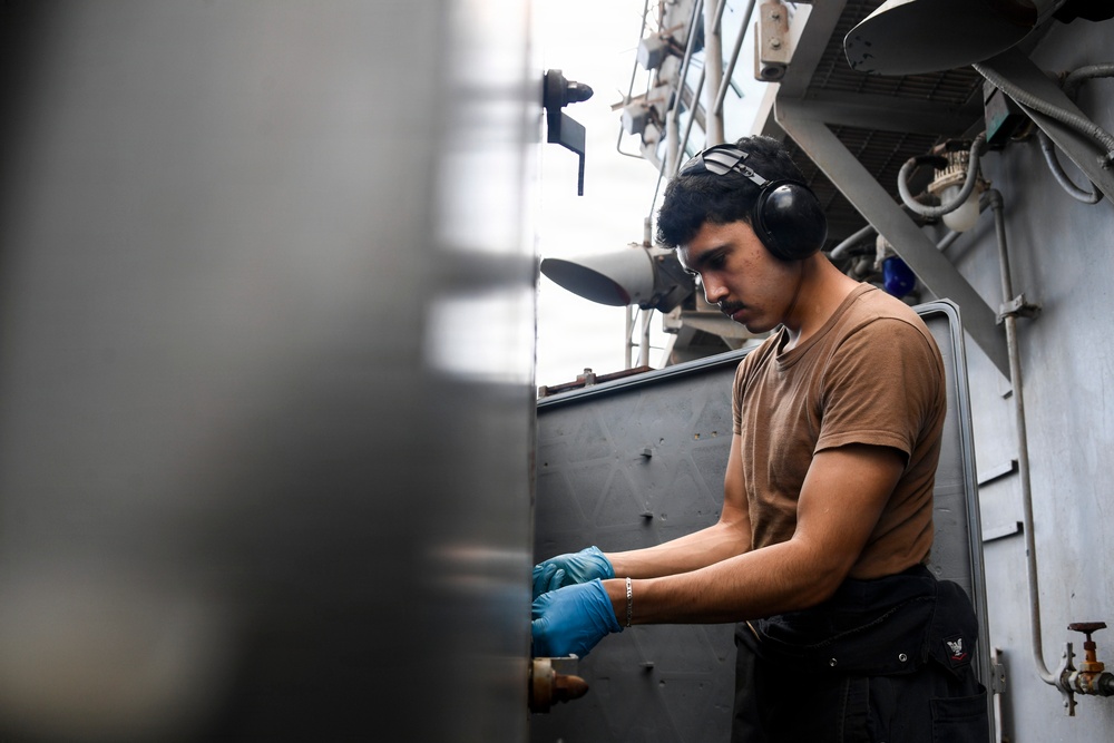 A young male worker is performing maintenance or mechanical work in what appears to be a naval vessel or industrial ship environment. He is wearing hearing protection (ear muffs) and blue nitrile gloves while working on a fitting or valve component. The setting shows a confined, metal-walled workspace with pipes, cables, and structural elements typical of a military or commercial ship.