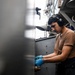 A young male worker is performing maintenance or mechanical work in what appears to be a naval vessel or industrial ship environment. He is wearing hearing protection (ear muffs) and blue nitrile gloves while working on a fitting or valve component. The setting shows a confined, metal-walled workspace with pipes, cables, and structural elements typical of a military or commercial ship.